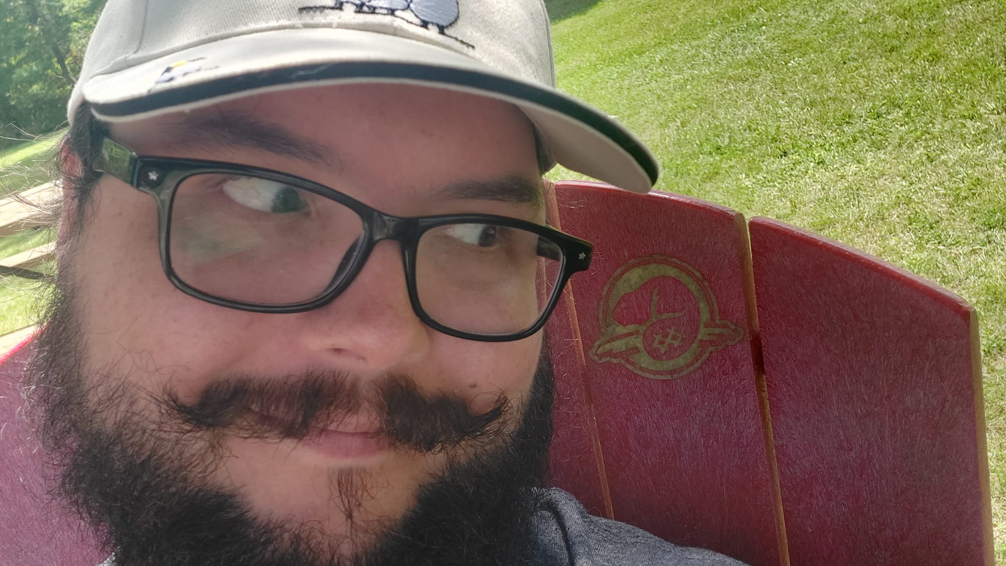 A man with short hair and bushy facial hair sits in a Muskoka chair, side eyeing a Parks Canada beaver logo stamped on it